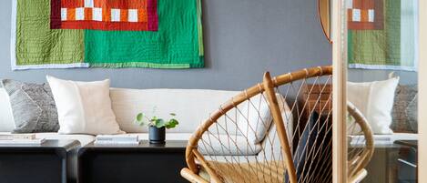 A modern living room with a wooden floor, a wicker chair, a sofa with cushions, and a large quilt hanging on the wall.