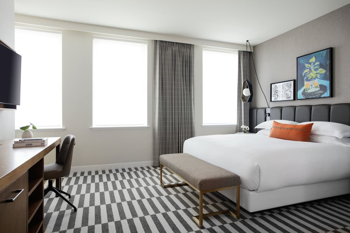 A hotel room with a large bed, a desk with a chair, a bench, and two windows with curtains.