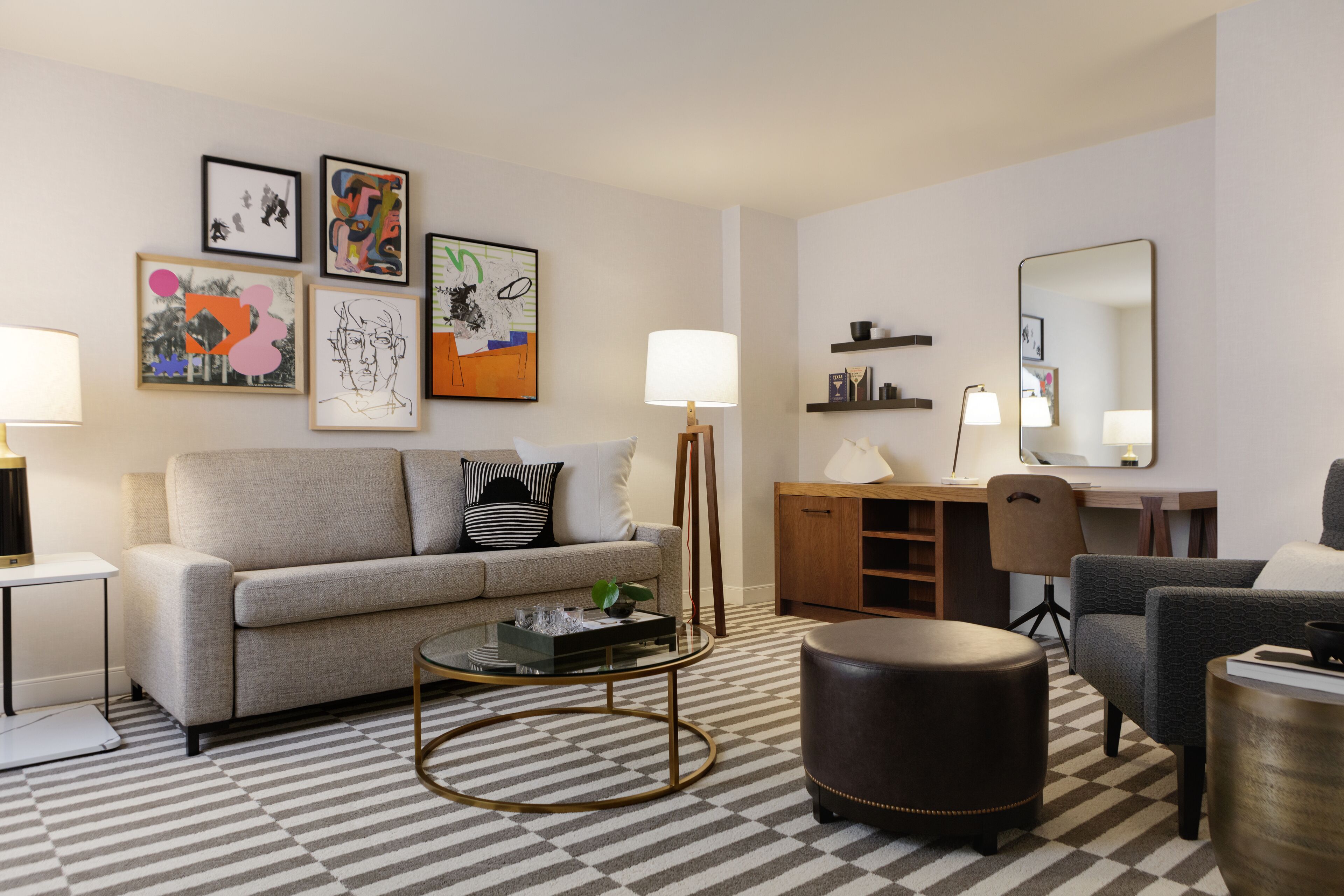 Un salon moderne comprenant un canapé, une table basse, un pouf et un bureau avec une chaise. Des œuvres d’art encadrées ornent le mur, et une lampe est posée sur le bureau.