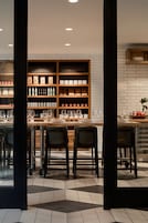 Una zona de bar con pared de ladrillo, estantes con botellas, vasos y una mesa con sillas.