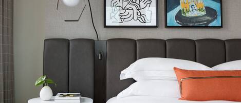 A modern bedroom with a large bed, a round wooden side table, a white vase with a plant, and two framed artworks on the wall.