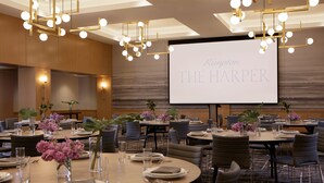 A conference room with multiple tables set for dining, equipped with chairs, plates, cutlery, and vases with flowers. A large screen displays the text "Kimpton THE HARPER".
