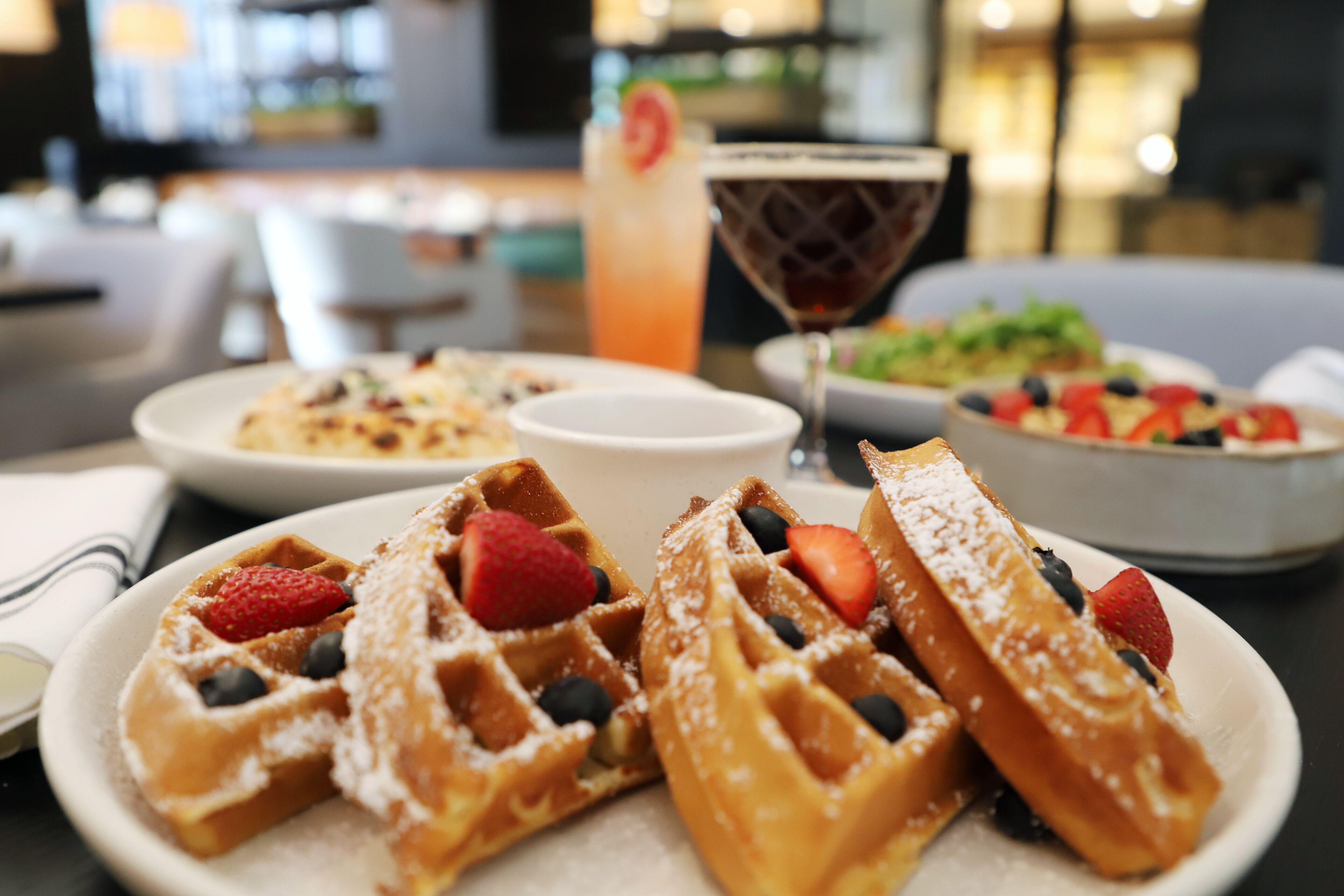 A table with waffles topped with strawberries and blueberries, a cocktail, and a bowl of salad.