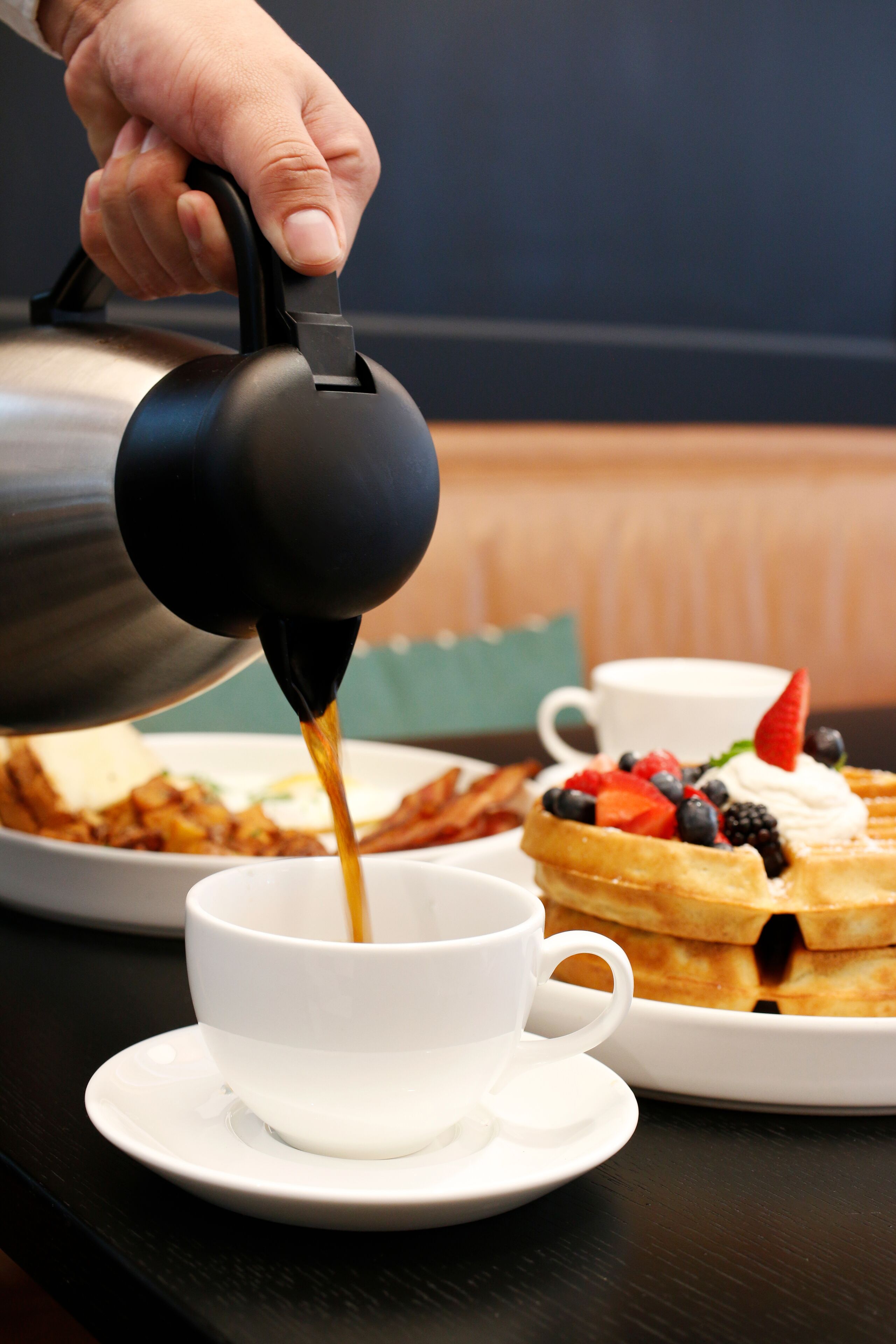 A hand pouring coffee into a cup, a plate of waffles with fruit, and a bowl of food on a table.