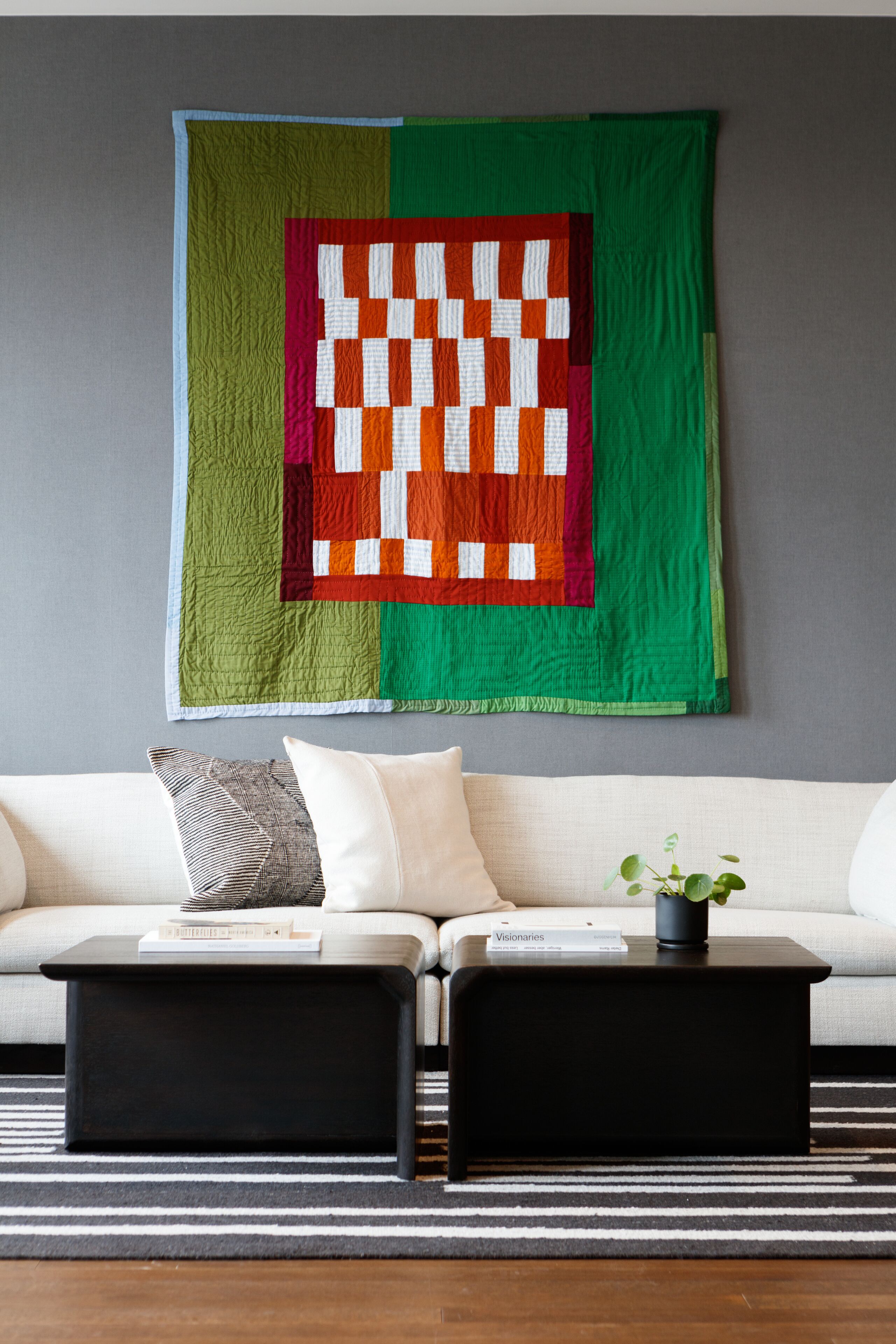 A living room with a large quilt hanging on the wall, a white sofa, two black side tables, and a striped rug.
