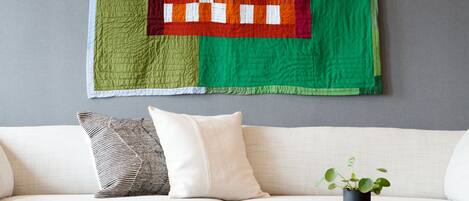 A living room with a large quilt hanging on the wall, a white sofa, two black side tables, and a striped rug.
