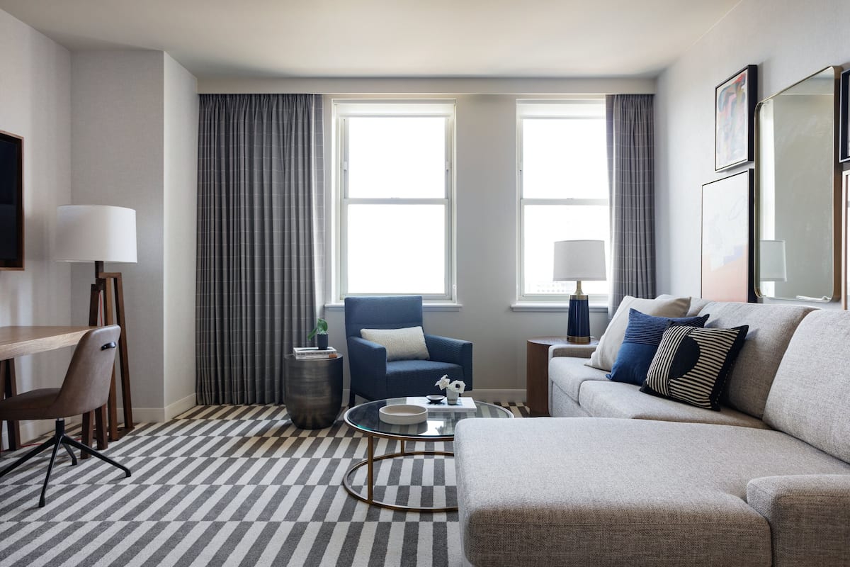 A modern hotel room with a checkered floor, a sofa, a chair, a coffee table, and two windows with curtains.