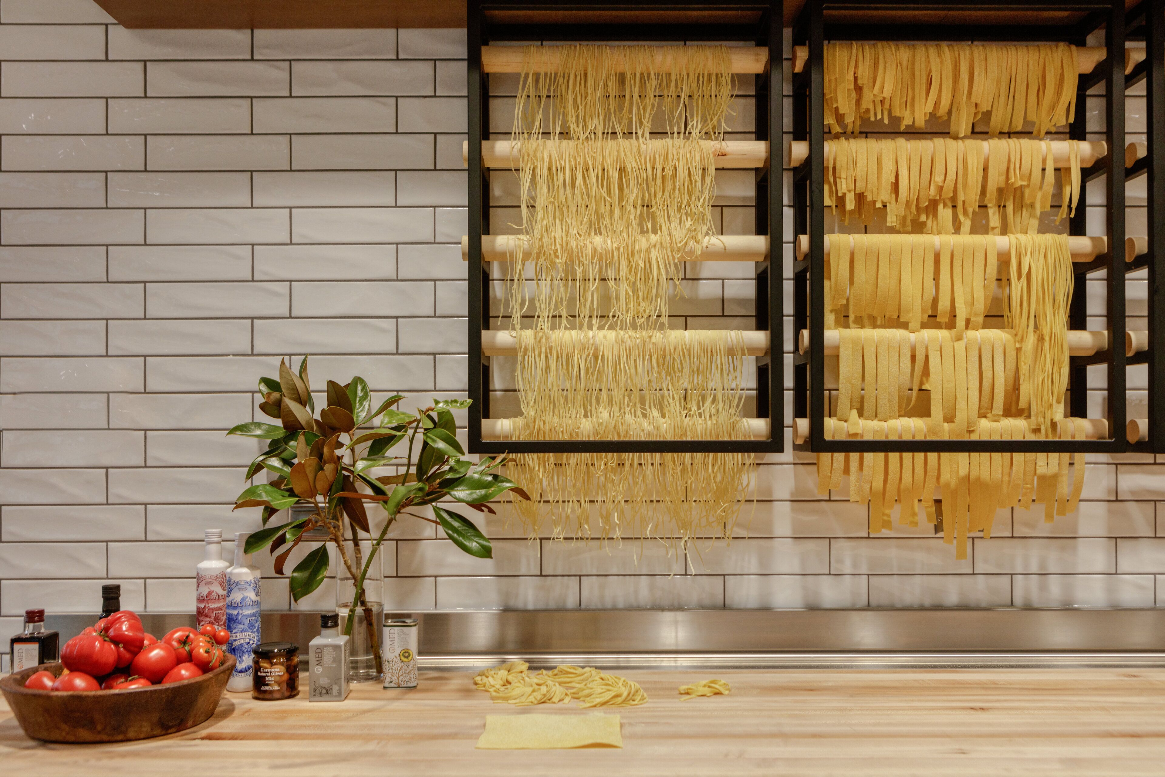 A kitchen with hanging pasta making equipment and a wooden bowl of tomatoes on the counter.