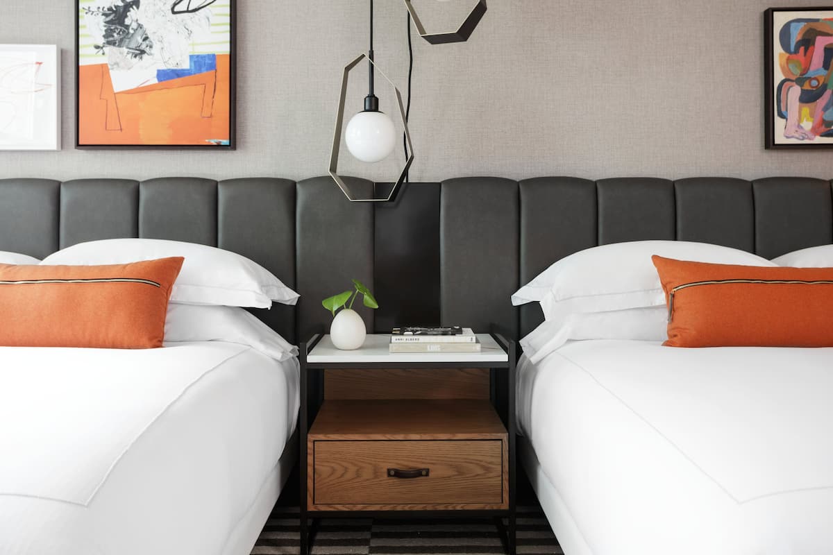 A hotel room with two beds, a nightstand with a plant, a book, and a wooden drawer.