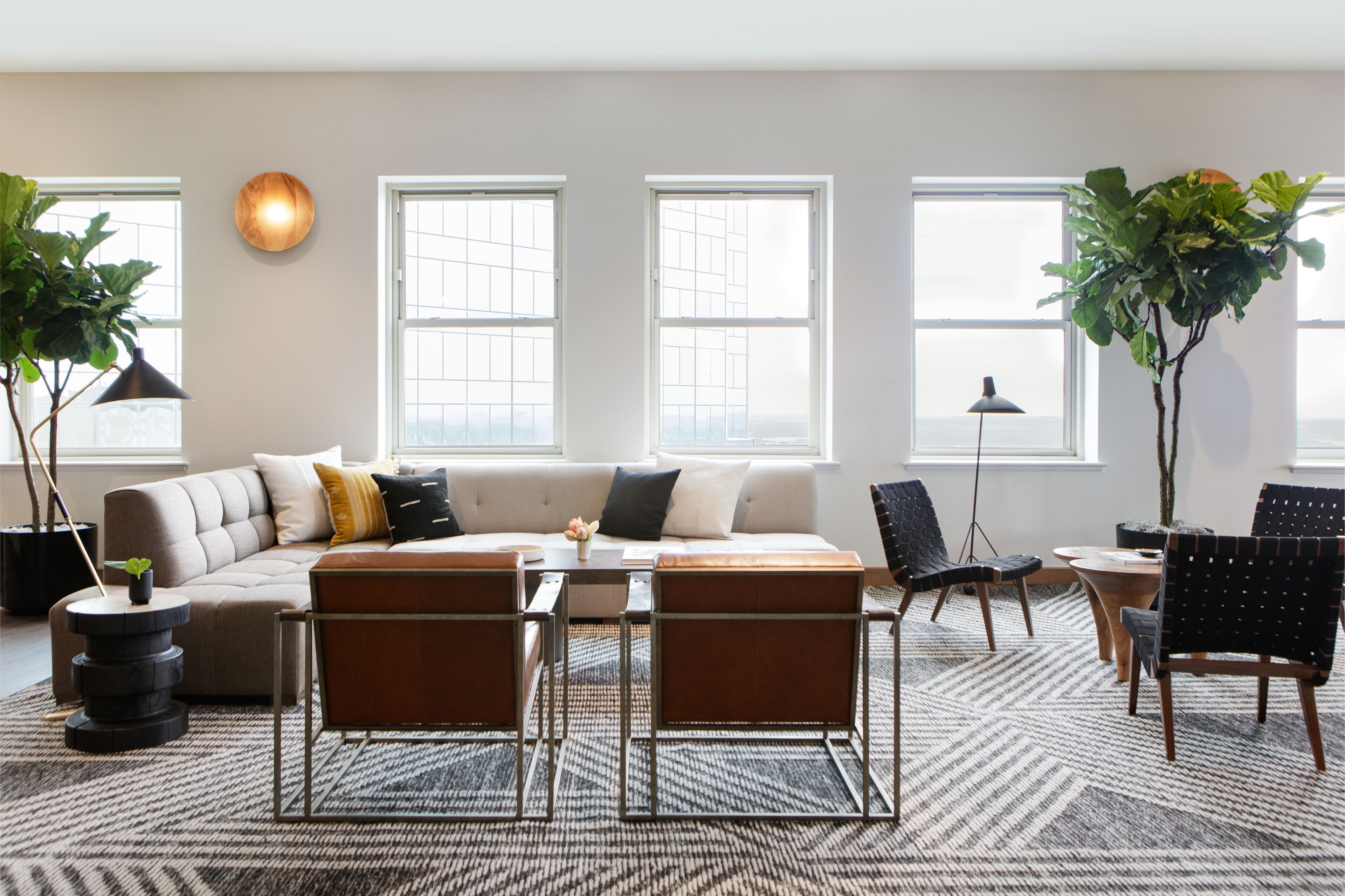 A modern living room with a sectional sofa, a coffee table, and two armchairs. There are large windows providing natural light and a view of the city.