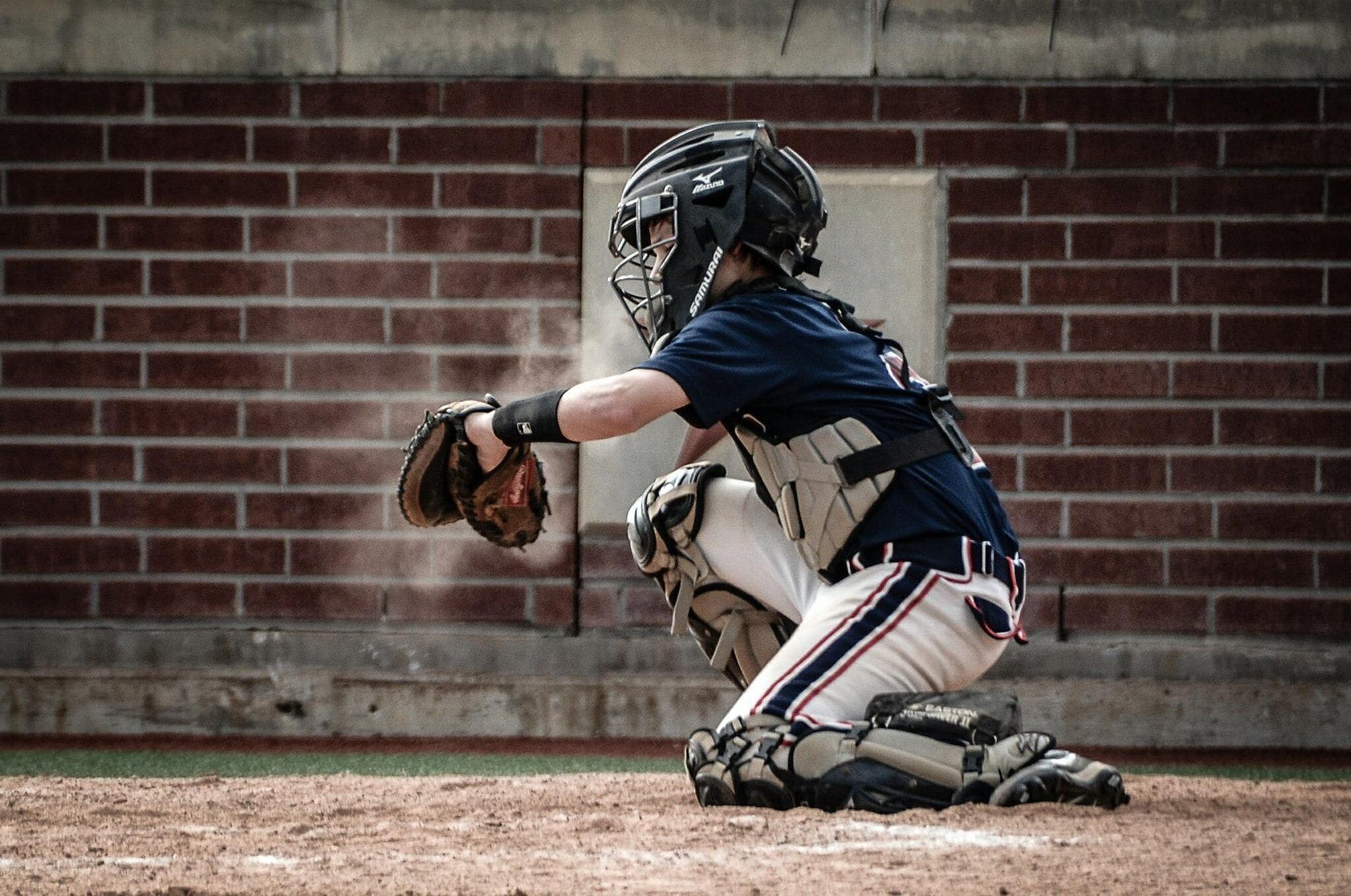 Um receptor de beisebol em posição de ataque, com luva de receptor, usando capacete e equipamentos de proteção, em frente a um muro de tijolos.