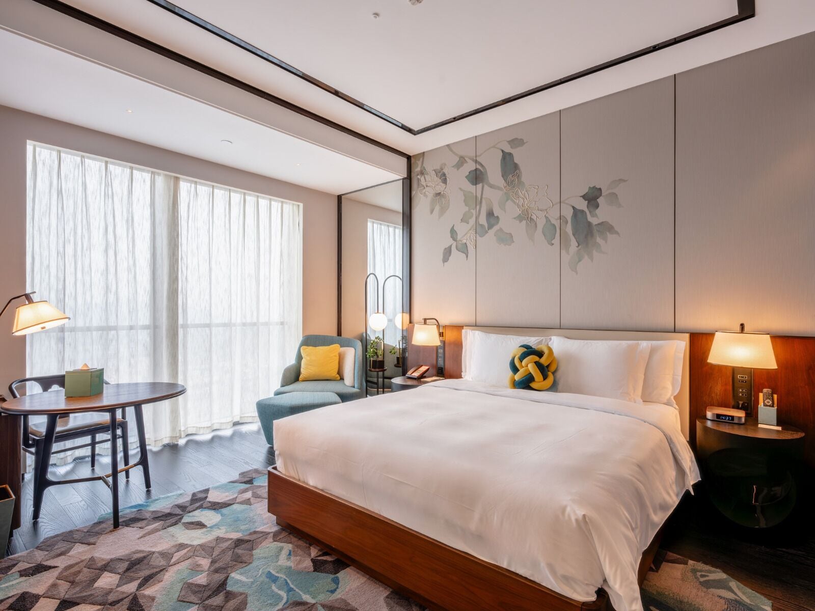 Une chambre d’hôtel avec un grand lit, un bureau avec une chaise, une petite table avec une lampe et une grande fenêtre avec des rideaux légers.