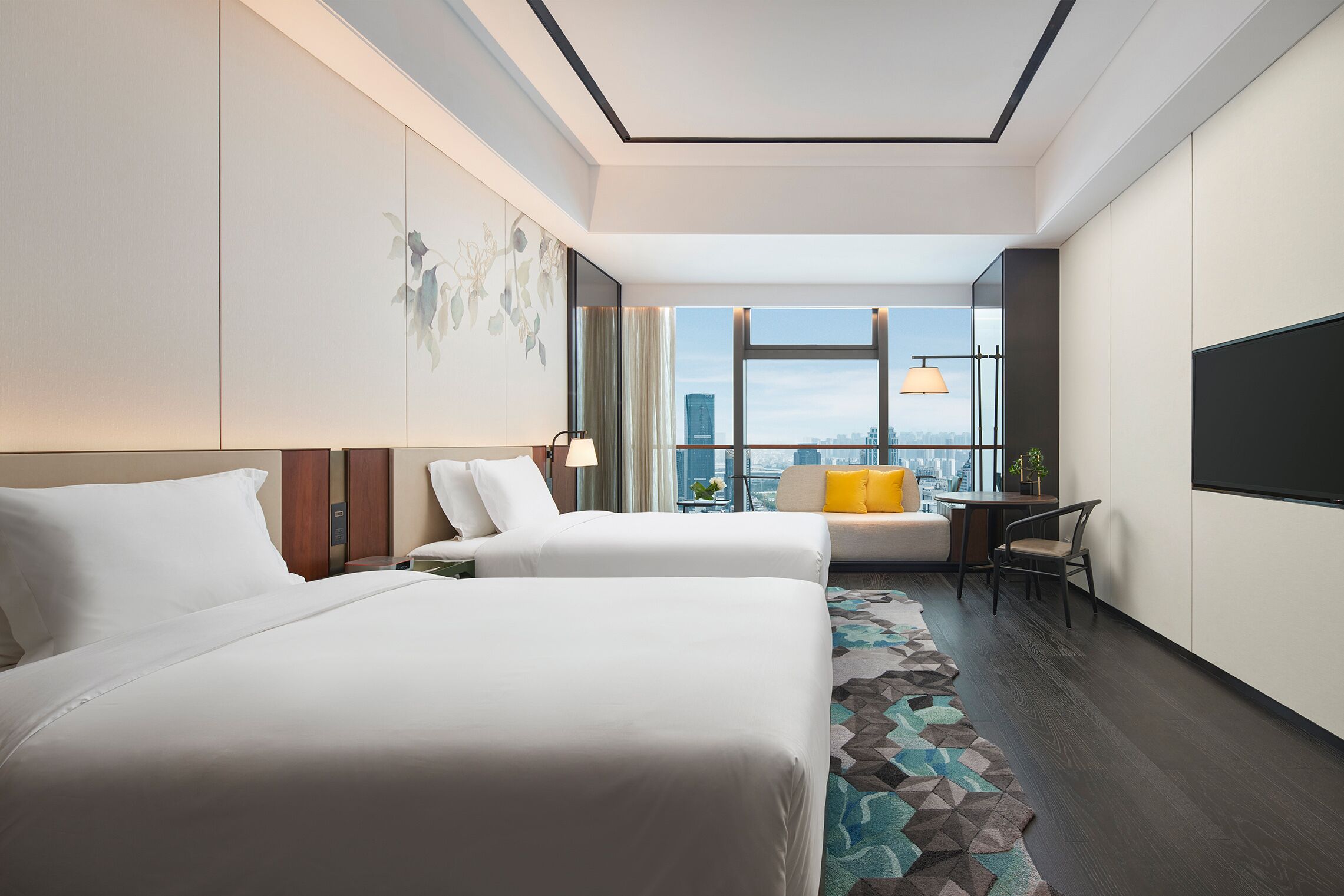 Habitación de hotel con dos camas, un escritorio, una silla, un ventanal amplio con vistas a la ciudad y una alfombra estampada.
