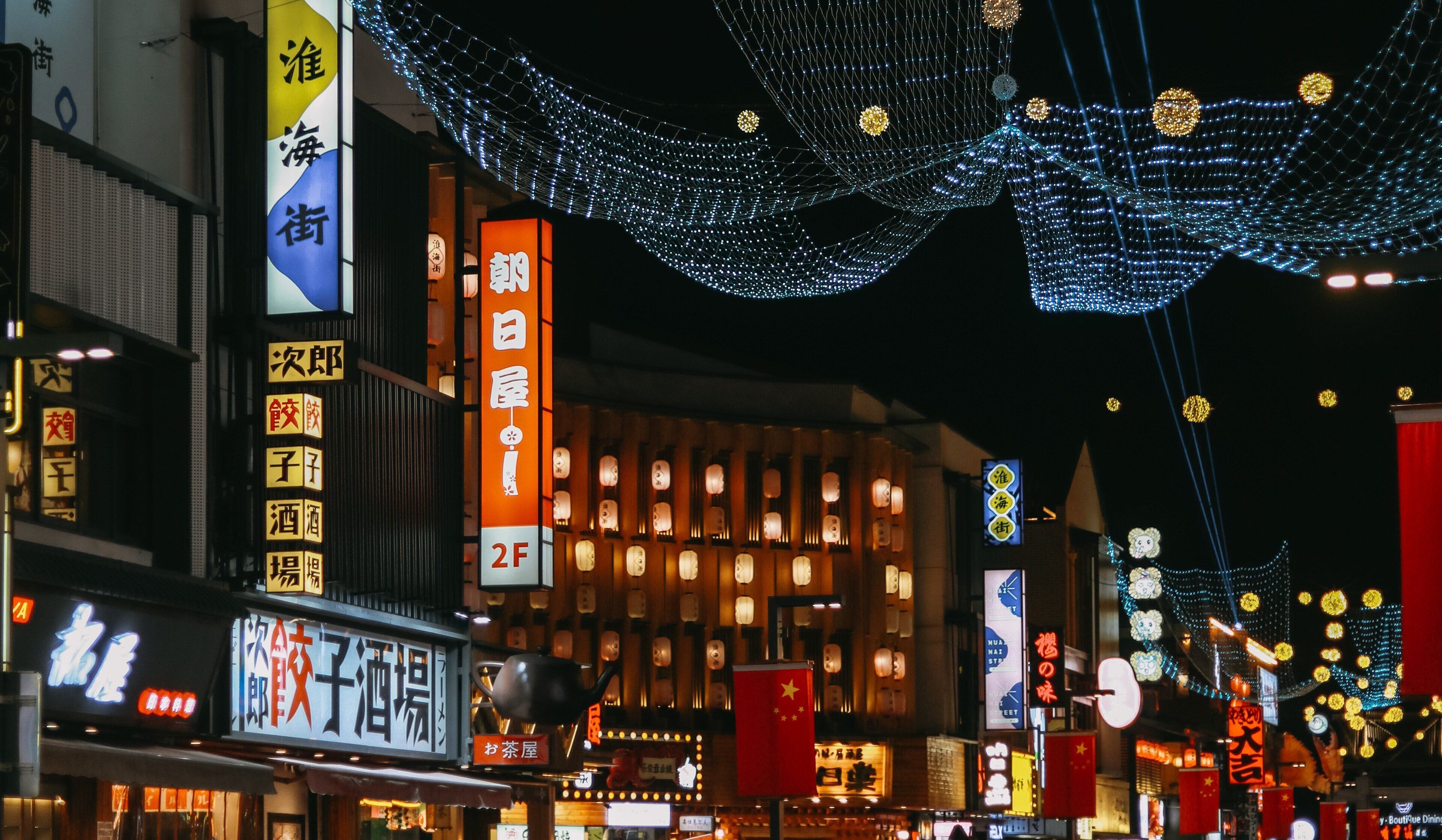 A street with illuminated signs and traditional lanterns, indicating a commercial area with shops and restaurants.