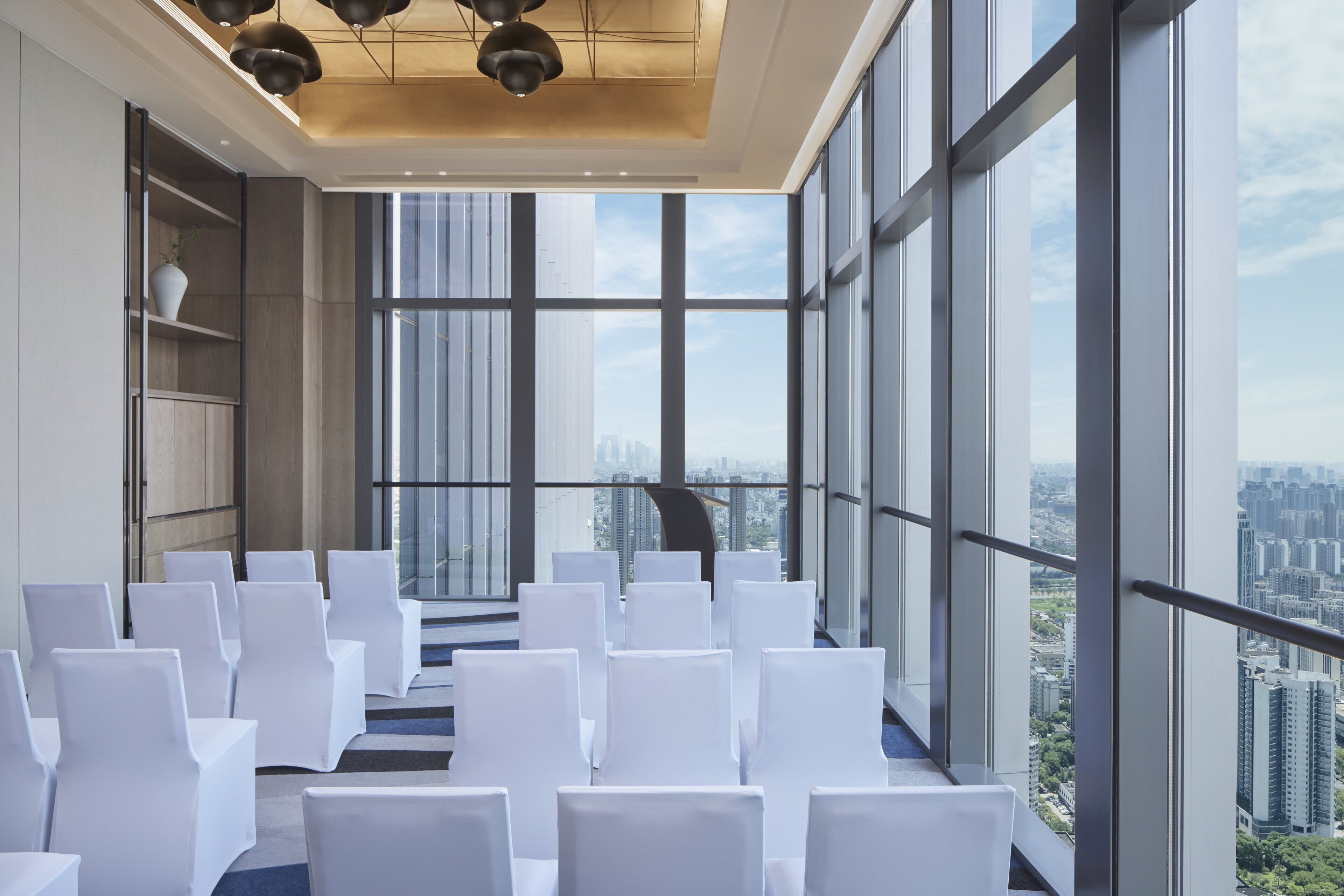Ein Konferenzraum mit weißen, in Reihen angeordneten Stühlen, großen Fenstern mit Blick auf die Stadt und einem modernen Deckenstil.