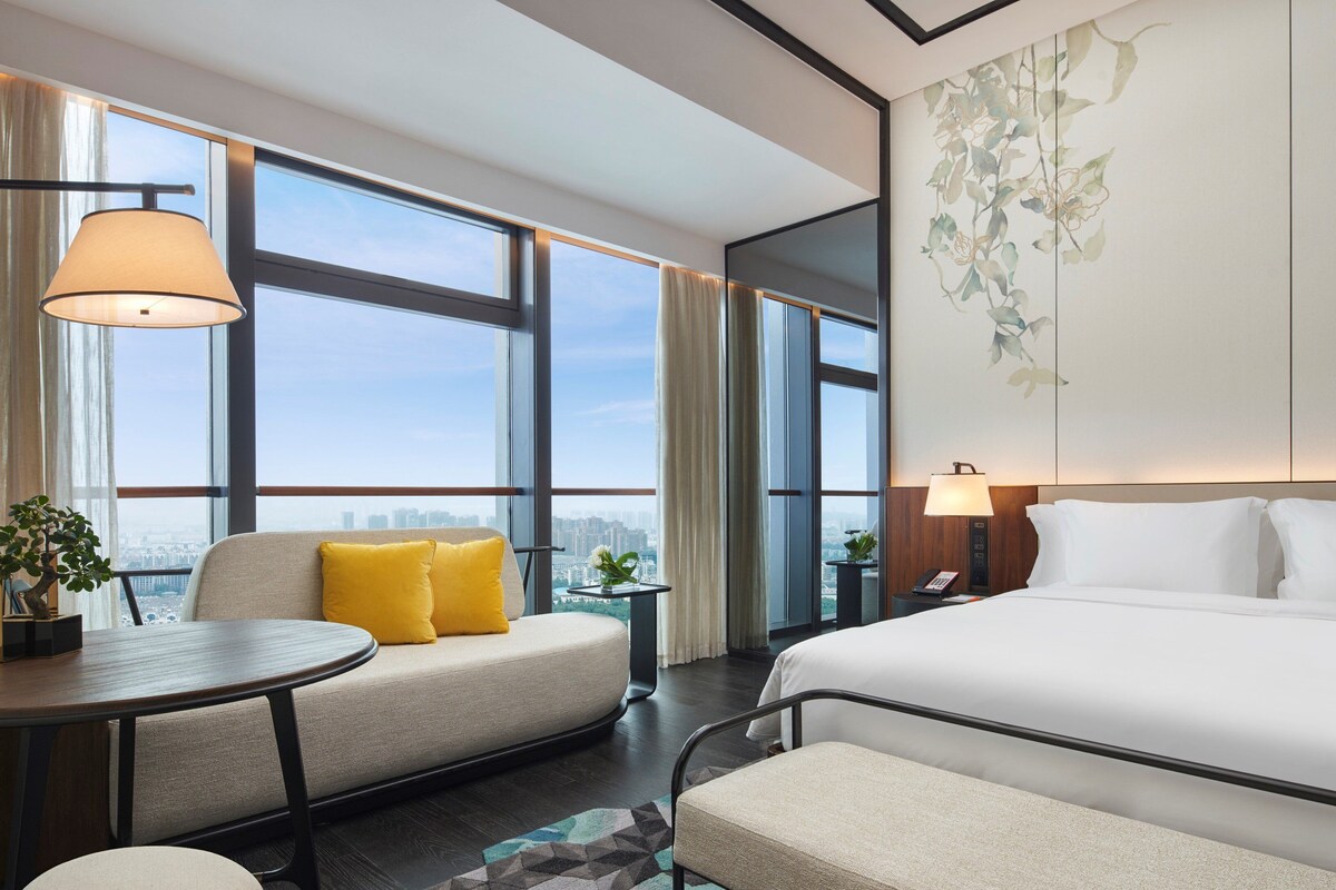 A hotel room with a large bed, a sofa, a small table, and a view of the city through large windows.