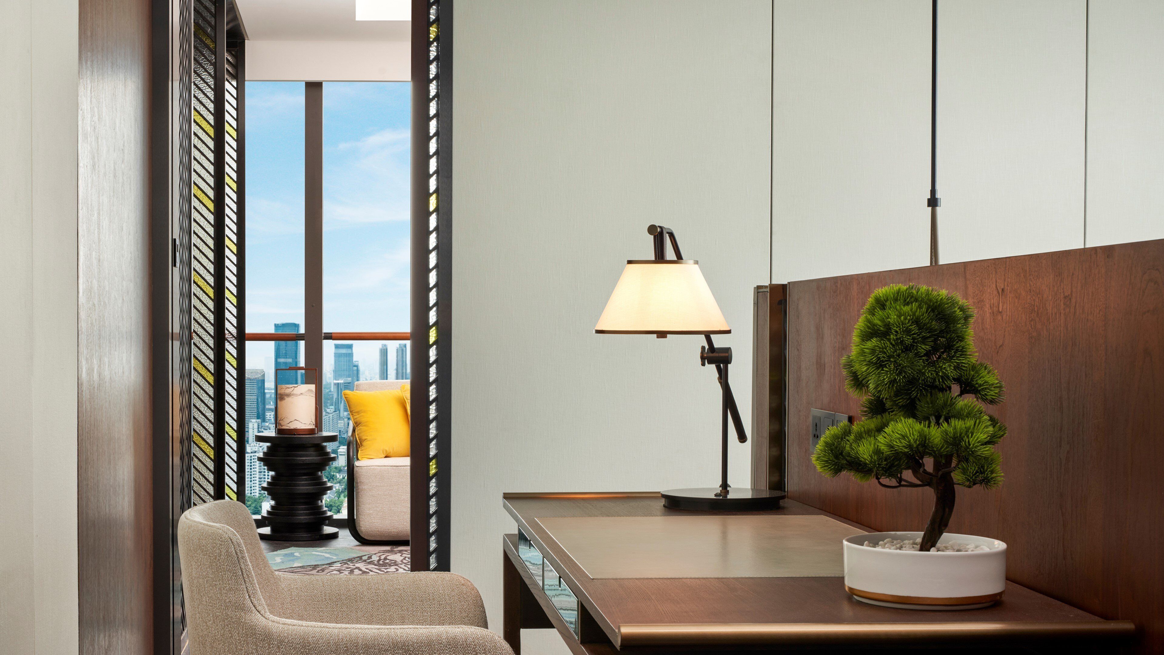 A modern hotel room with a city view, a desk, a lamp, a chair, and a bonsai tree.