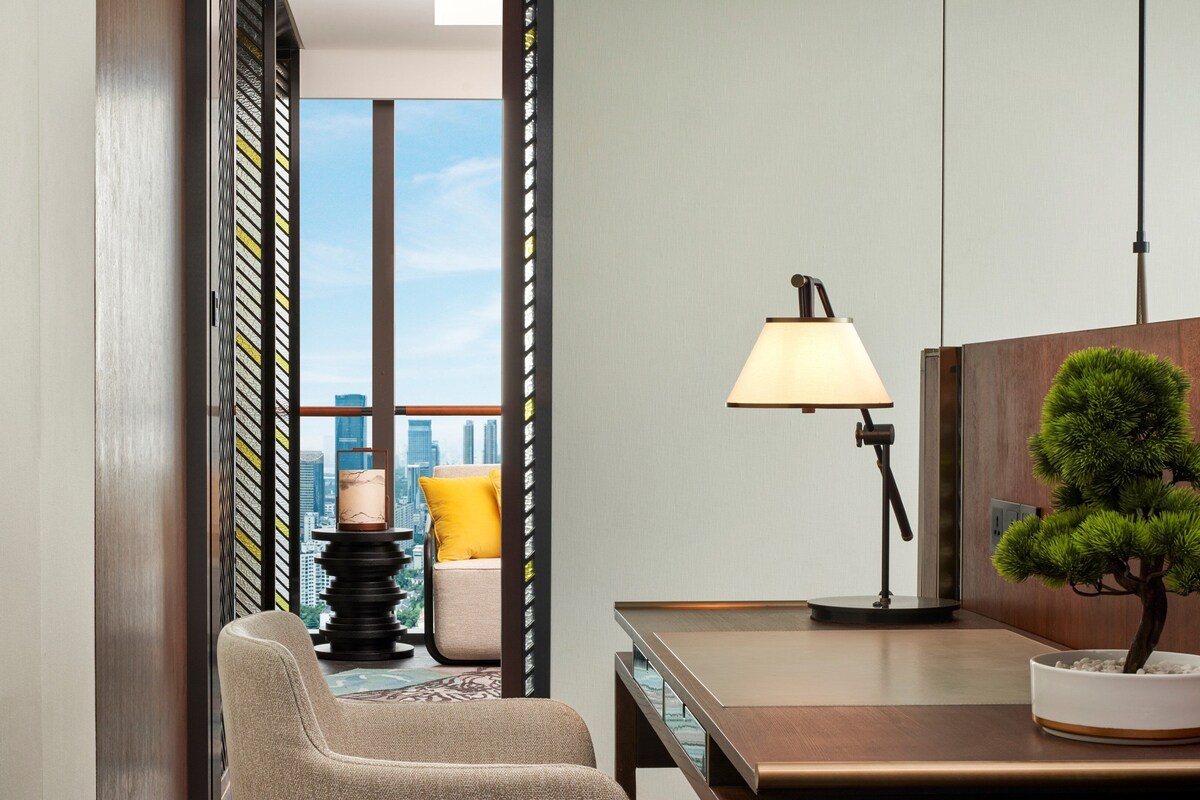 A modern hotel room with a city view, a wooden desk, a lamp, a chair, and a bonsai tree.