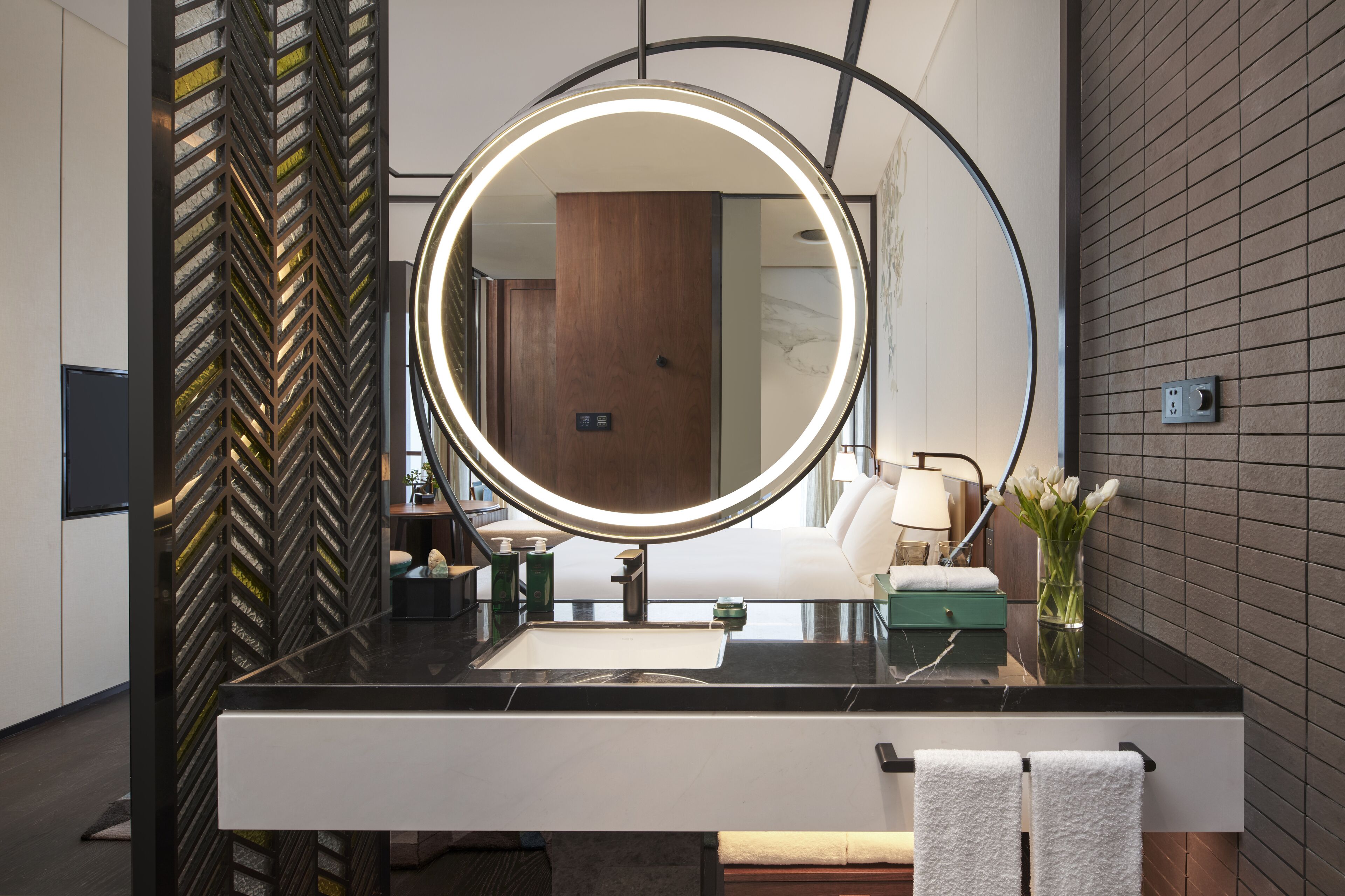 A modern bathroom with a black marble countertop, a large mirror with a circular light, a wooden door, and a vase with flowers.