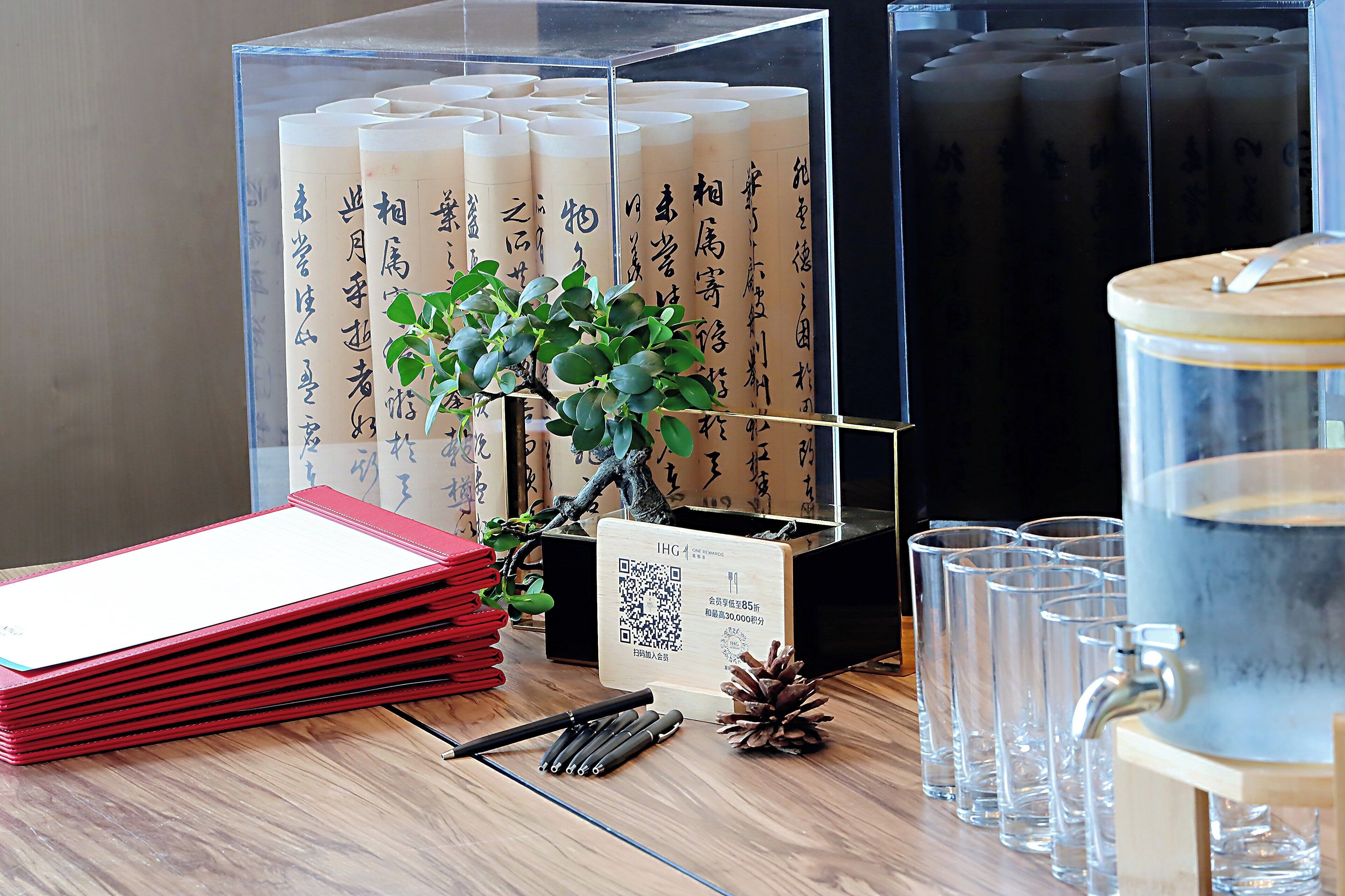 A wooden table with a stack of red folders, a QR code sign, a water dispenser, and several clear glasses.