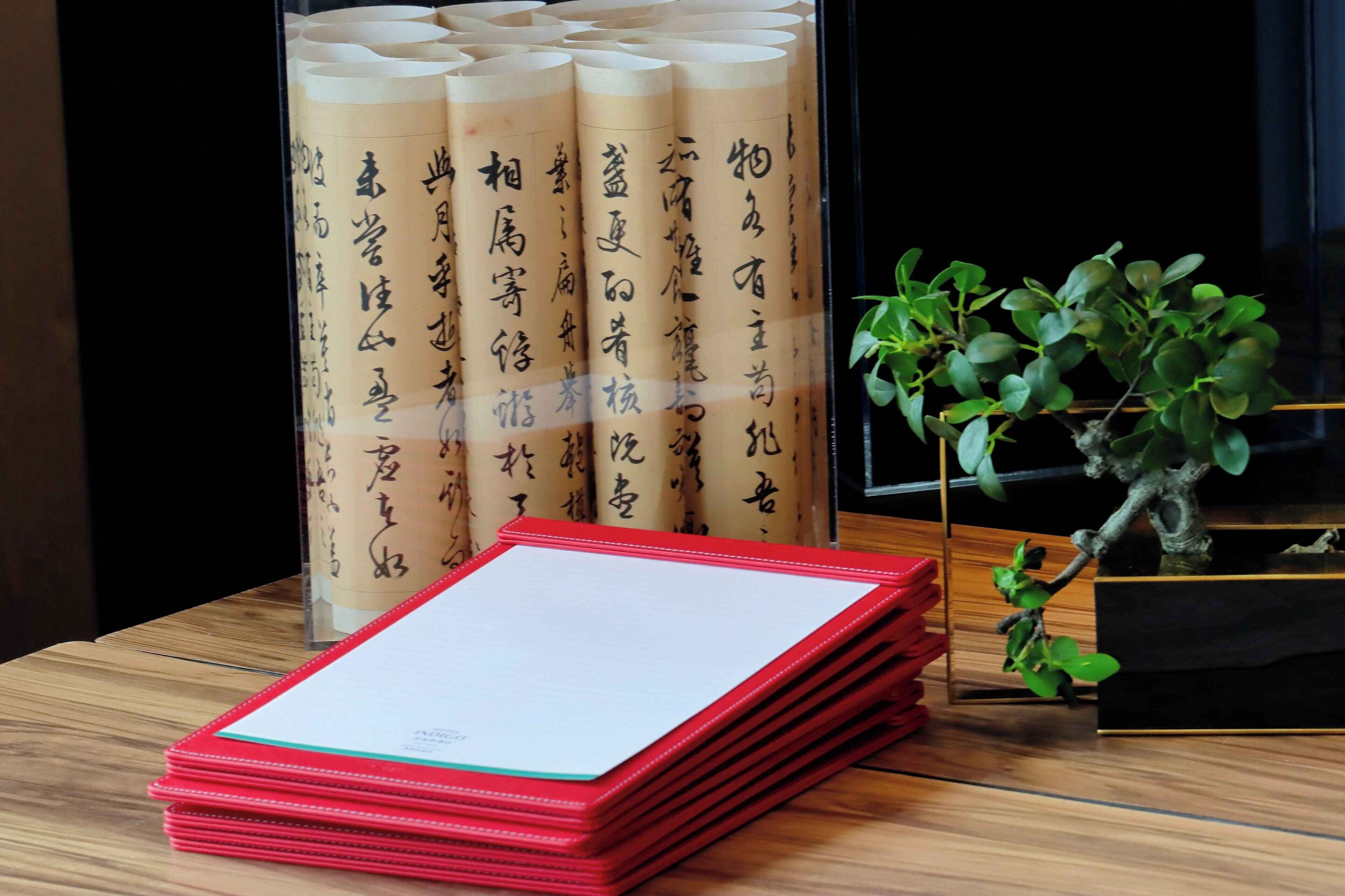 Una pila de carpetas rojas sobre una mesa de madera, un expositor de vidrio con documentos enrollados y una planta en maceta.