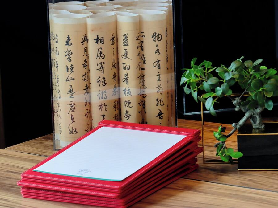 Una pila de carpetas rojas sobre una mesa de madera, un expositor de vidrio con documentos enrollados y una planta en maceta.
