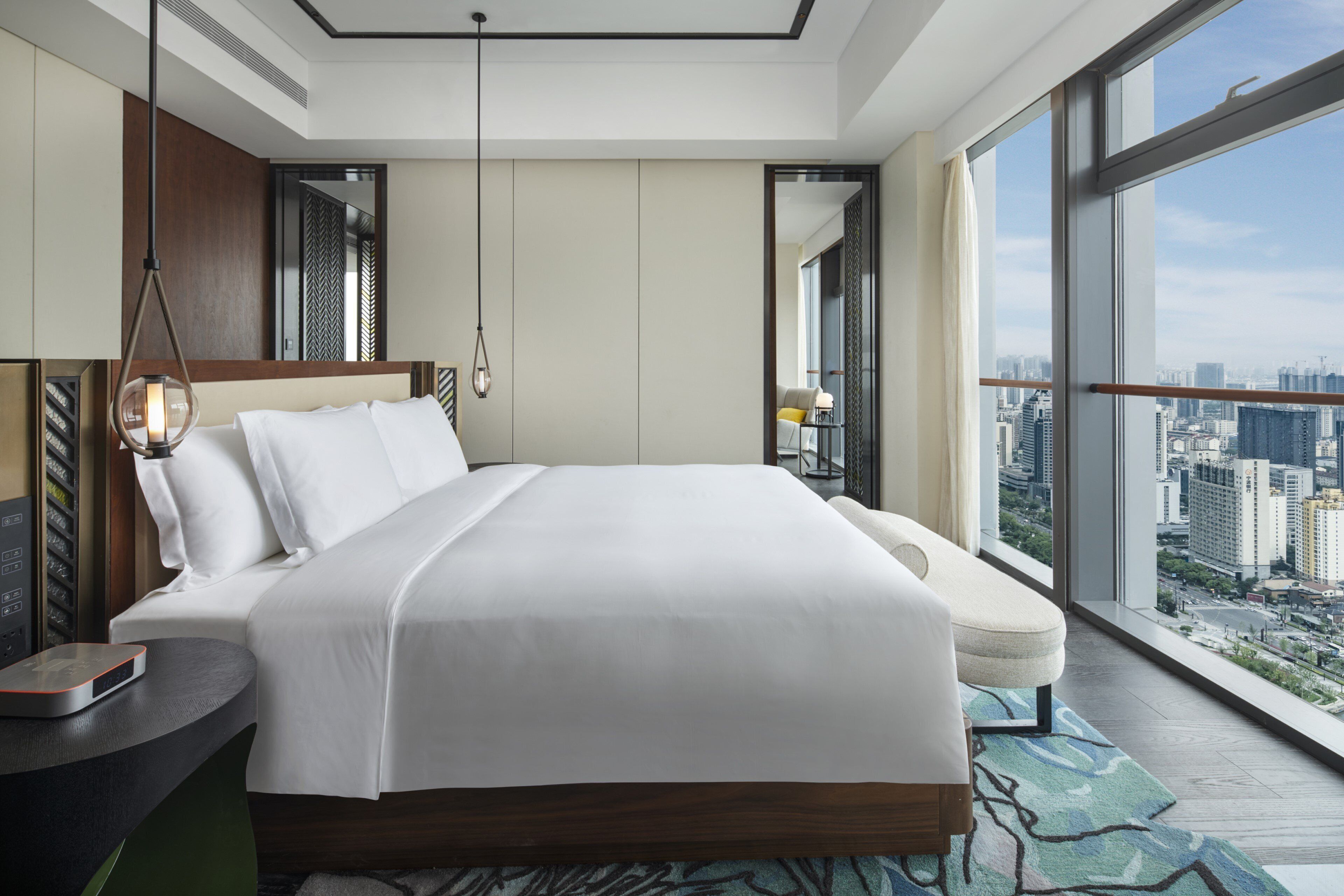 A modern hotel room with a large bed, a bedside table with a lamp, a view of the city through large windows, and a cityscape visible outside.