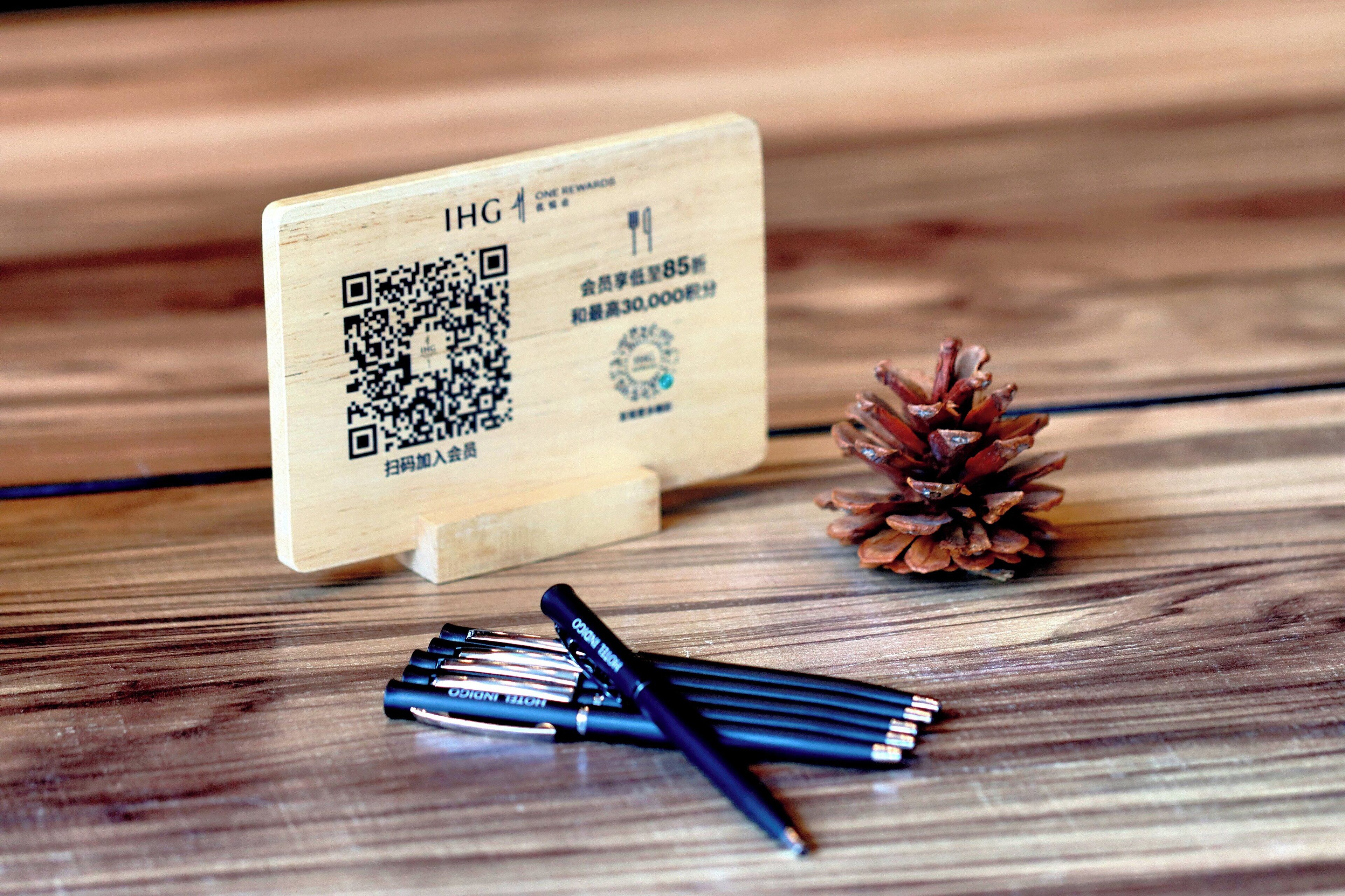 A wooden card with QR code and text, a pine cone, and pens on a wooden surface.