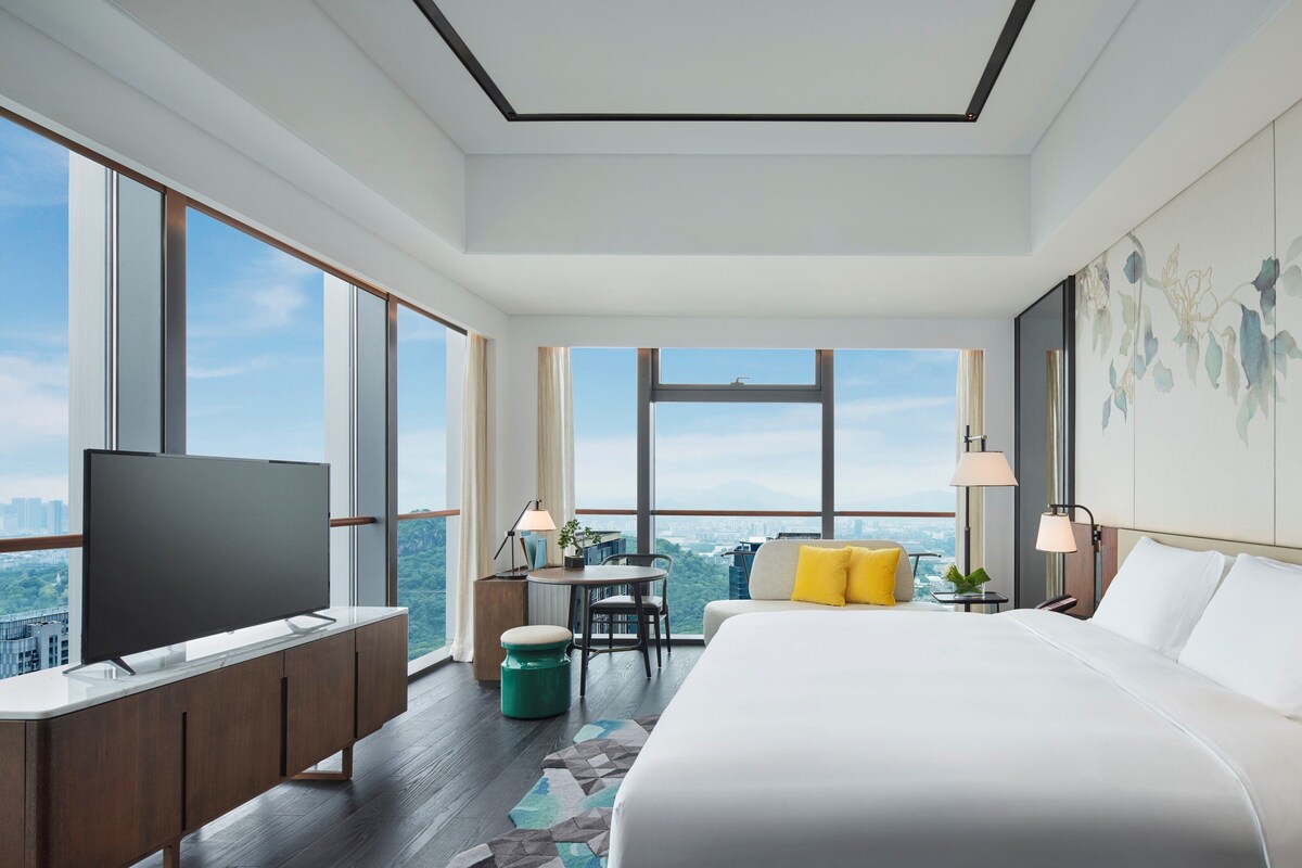 A modern hotel room with a large bed, a TV, a desk with a chair, and a view of the city.