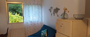 Una habitación con ventana, una cama con cubierta azul, un vinilo decorativo de un mapa mundial, un jarrón pequeño con flores y un radiador.
