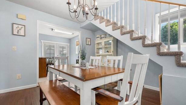 Un coin repas avec une table en bois et des chaises blanches, un escalier avec des rampes blanches et un lustre.