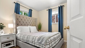 Une chambre à coucher avec une tête de lit capitonnée, un lit recouvert d’une couverture à carreaux, une table de chevet avec une lampe et une fenêtre ornée de rideaux bleus.