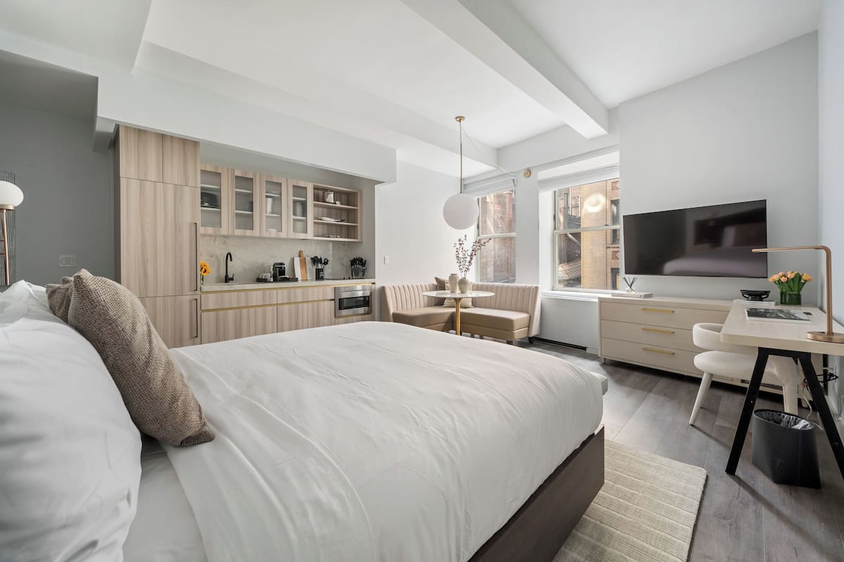 A modern bedroom with a large bed, a desk, a TV, and a kitchen area.