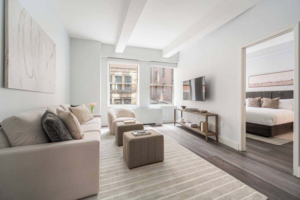 A modern bedroom with a bed, a sofa, a coffee table, a TV, and a window with a view of buildings.