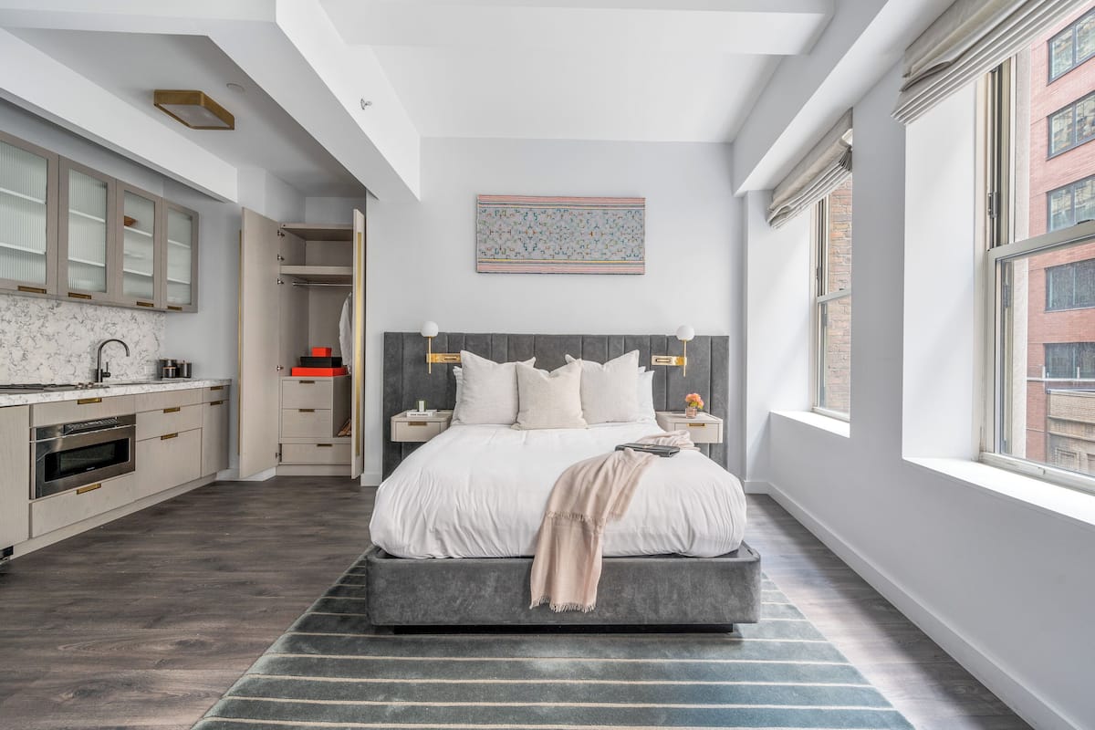 A modern bedroom with a large bed, a built-in wardrobe, a bathroom with a shower, and a view of the city through large windows.