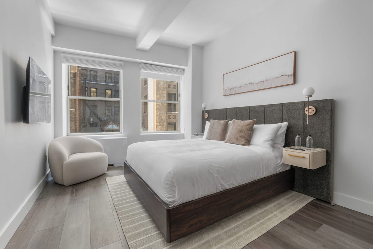 A modern bedroom with a large bed, a bedside table, a chair, and a window with a city view.