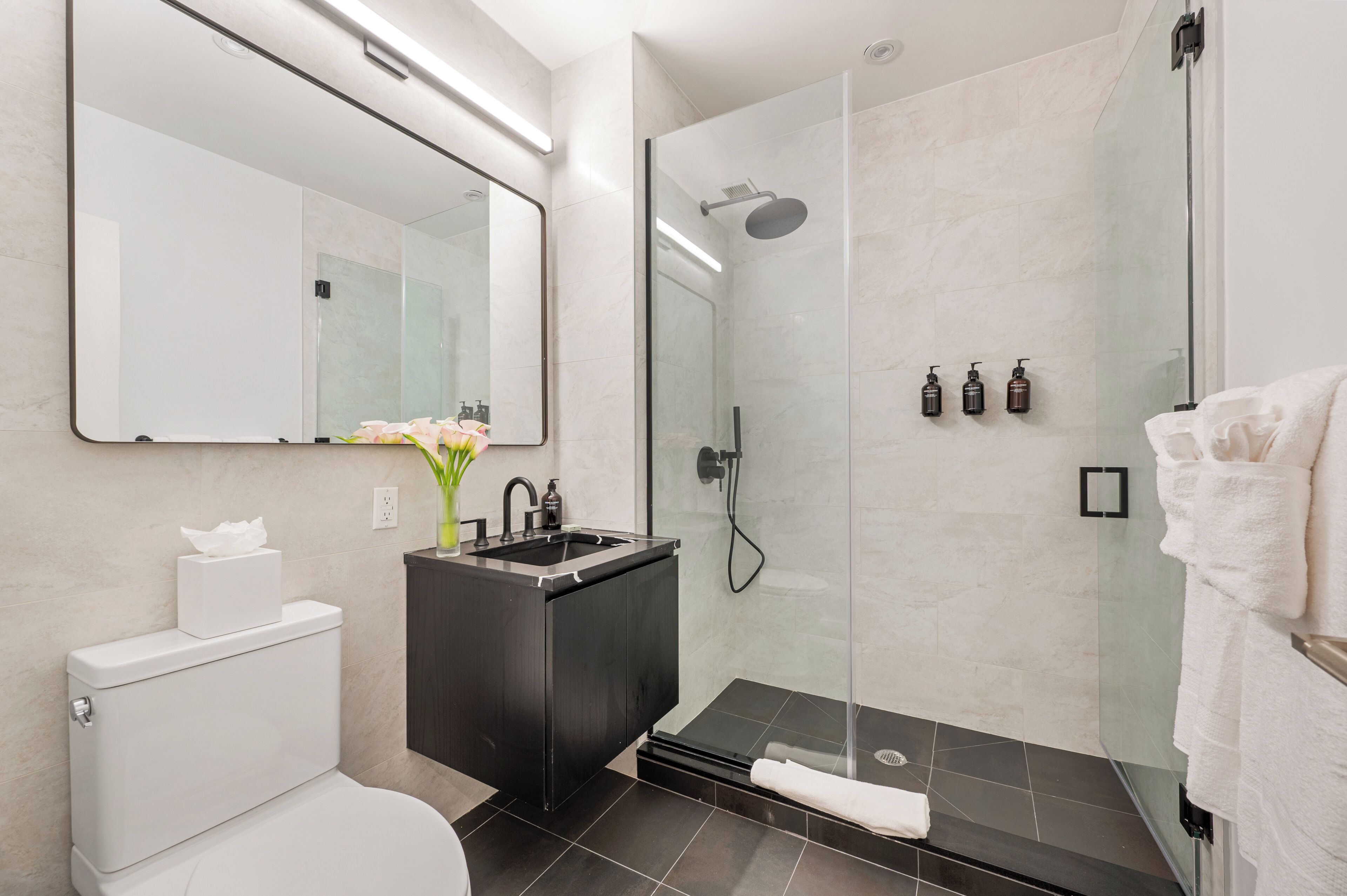 A modern bathroom with a glass-enclosed shower, a black vanity, a toilet, and a large mirror.
