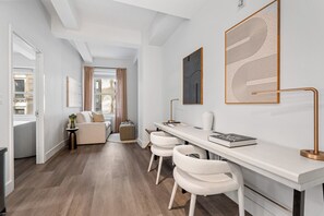 A modern hotel room with a long white desk, two white chairs, a bed, a small side table, and artwork on the walls.