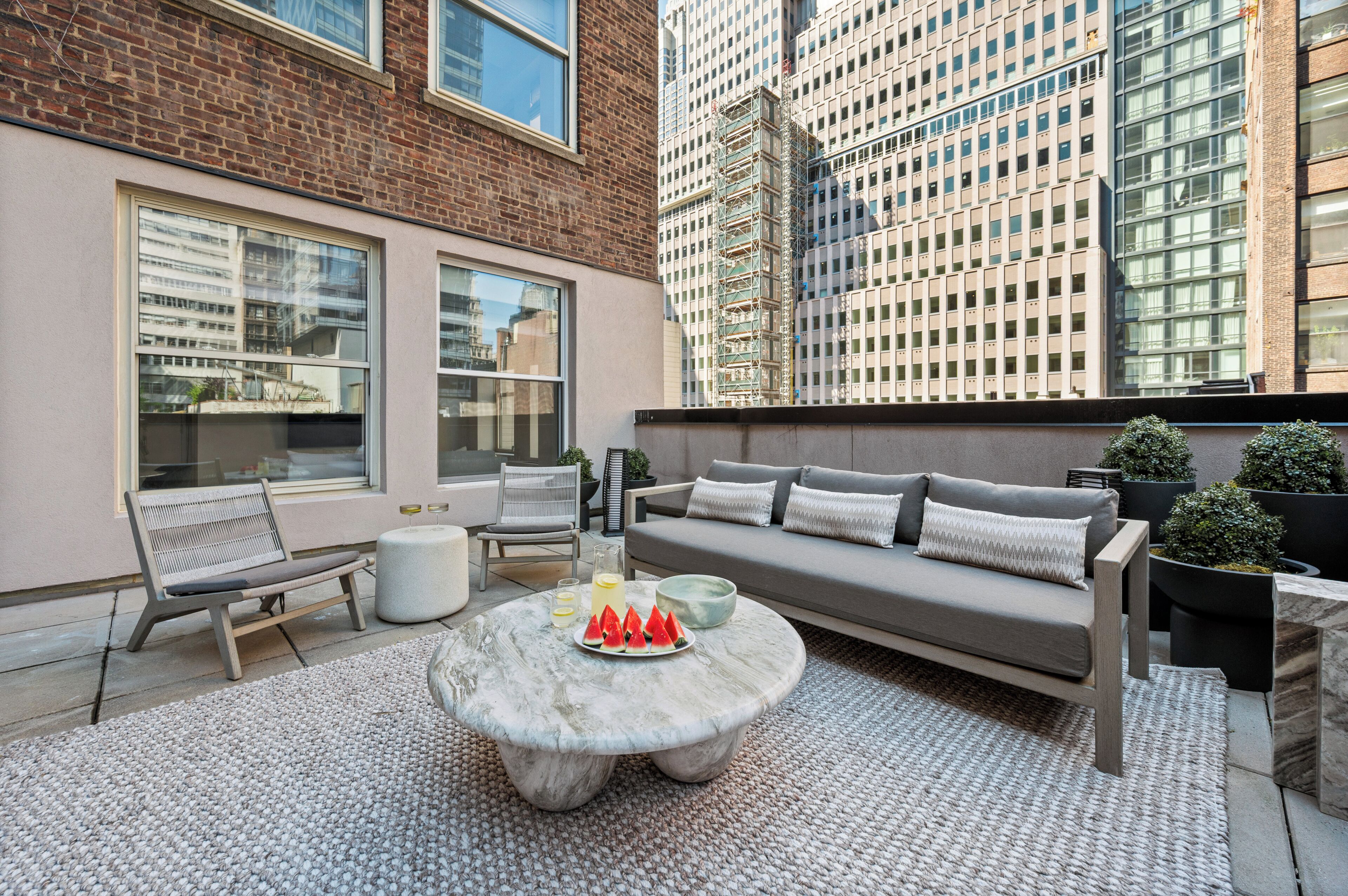 A rooftop patio with a sofa, chairs, a round table with a fruit platter, and a view of tall buildings.