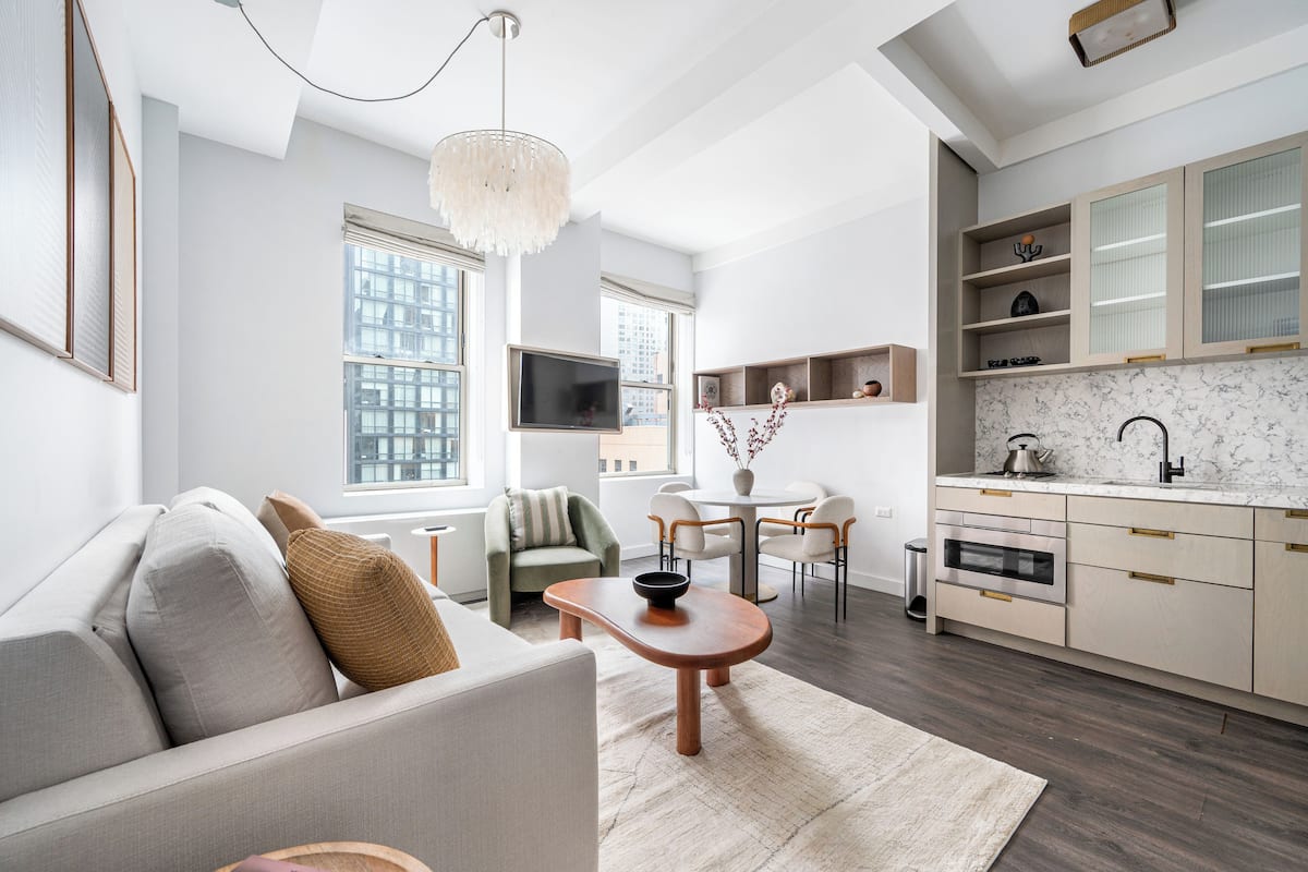 A modern living room with a sofa, coffee table, and TV. There is a kitchen with a dining area and a large window.