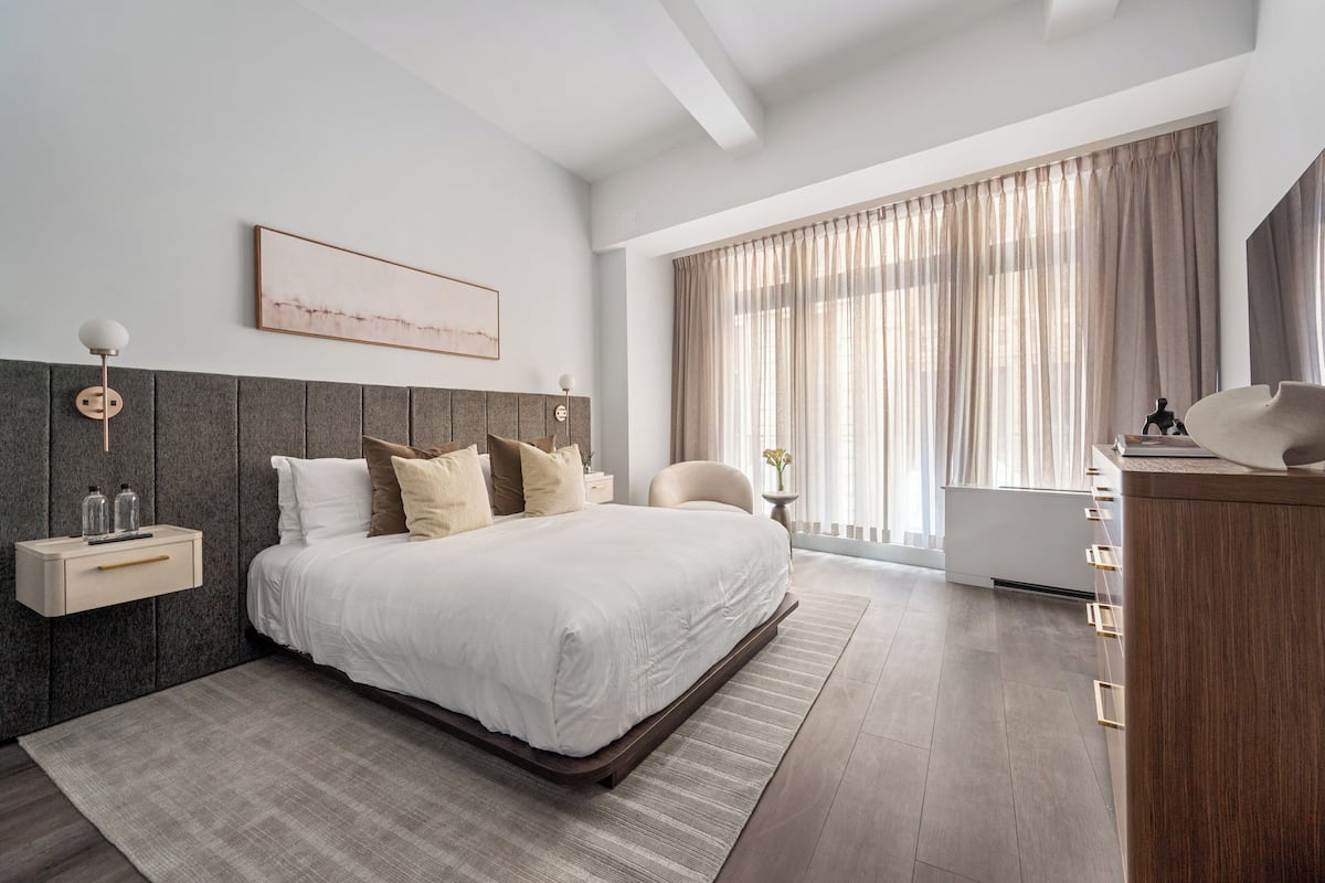 A modern bedroom with a large bed, a bedside table with a lamp, a desk, and a large window with curtains.