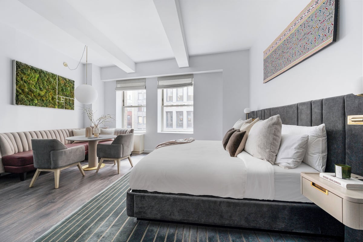 A modern hotel room with a large bed, a sofa, a dining area, and a view of the city.