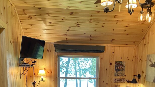 A cozy cabin interior with a wooden ceiling, a large window, a television mounted on the wall, and a fireplace. - Escape to cozy, secluded lakefront cabin in the north woods (Squaw Lake)