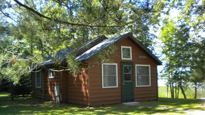 A small wooden cabin with a green door, two windows, and a small porch area. - Escape to cozy, secluded lakefront cabin in the north woods (Squaw Lake)