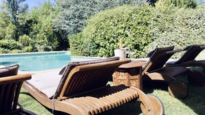 A poolside area with wooden lounge chairs and a table, surrounded by lush greenery and trees.