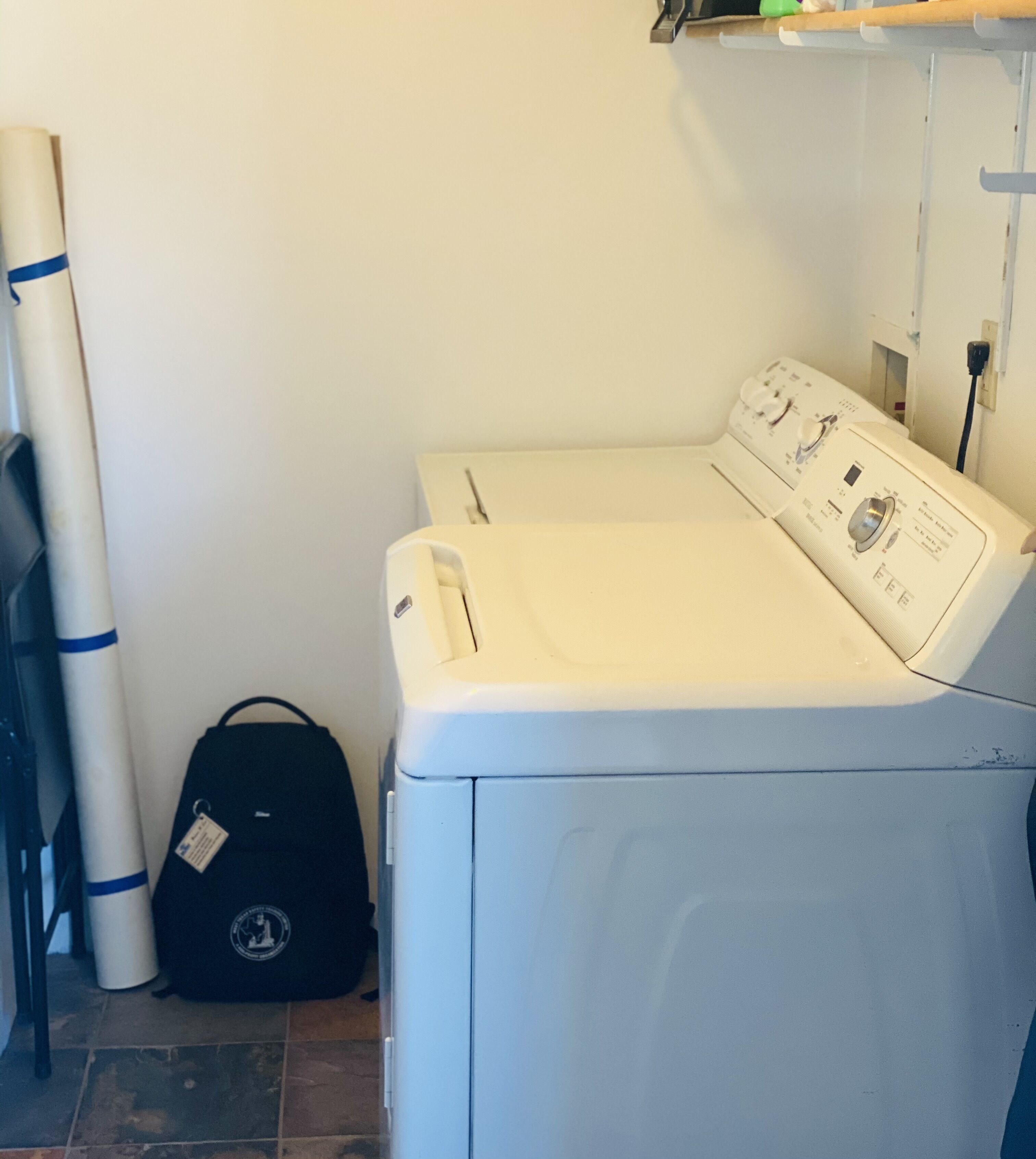 Een voorlader wasmachine met een bedieningspaneel, een zwarte tas en een witte, cilindervormige voorwerp tegen een effen muur.