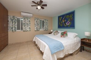 Un dormitorio con cama, ventilador de techo, aire acondicionado y un cuadro con temática tropical.