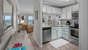 A modern kitchen with white cabinets, a dishwasher, a stove, and a microwave. There is a view of the ocean through a large window. - 2/2 Oceanfront Condo w/ Pool - Windy Hill (North Myrtle Beach)