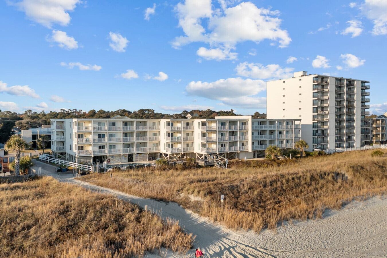 A beachfront hotel with multiple floors, balconies, and a sandy path leading to the entrance.