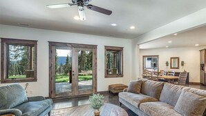 Un salon spacieux avec un grand canapé, une table basse et un ventilateur de plafond. De grandes fenêtres offrent une vue sur une zone boisée.